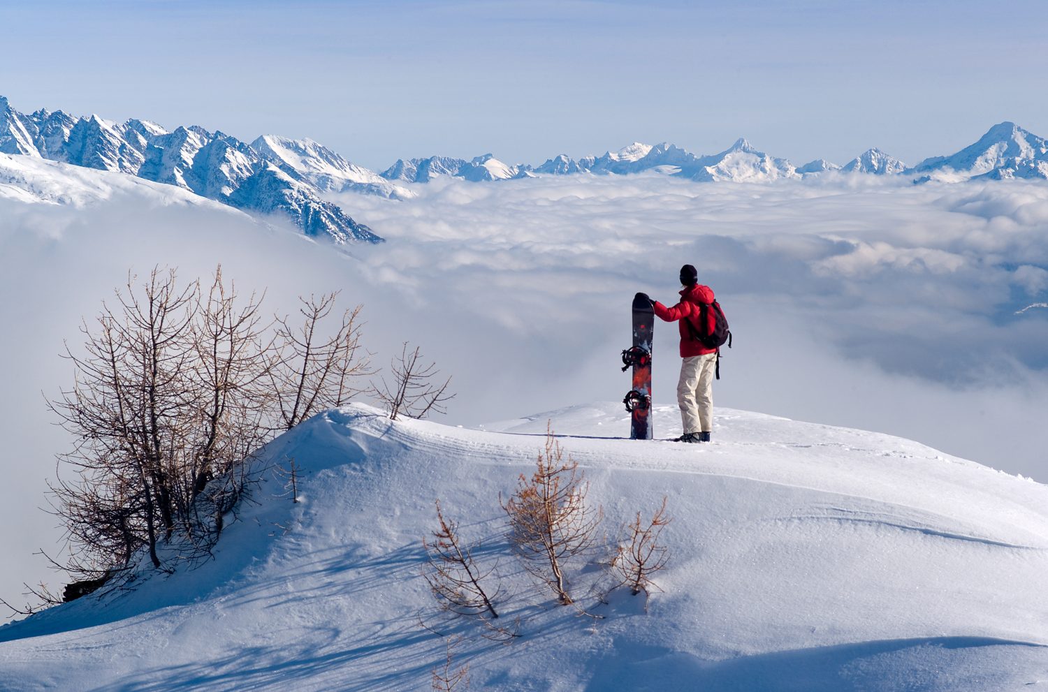 A snowboarder observes the panorama, with a cloud cover beneath him. Photo Olivier Maire- Courtesy: Crans-Montana Tourism Office.